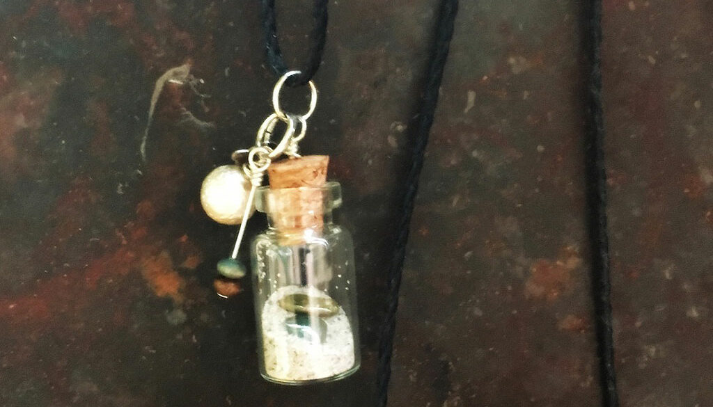 Hänge glasflaska innehållande sand och två små stenar. Små amuletter hänger från korken.